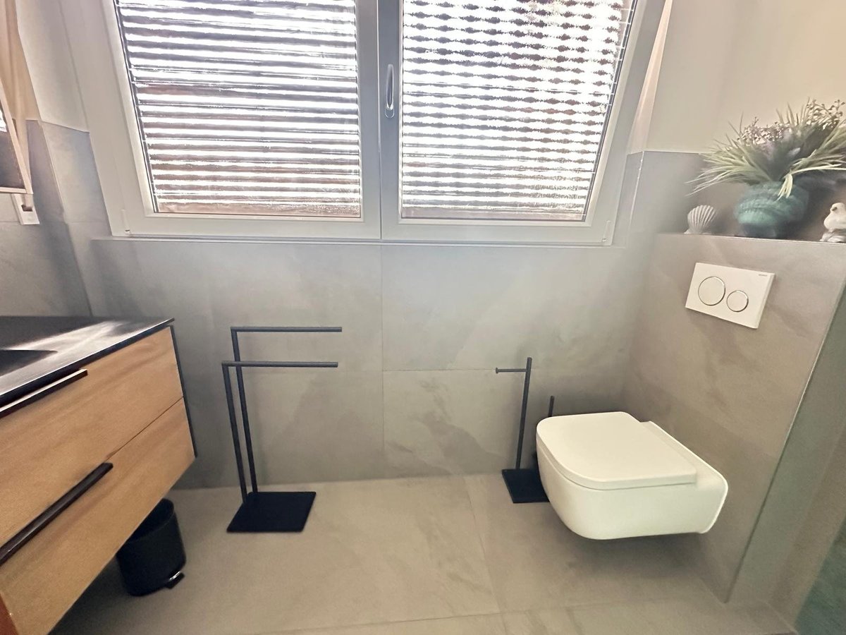Modern bathroom with wall-mounted white toilet, towel racks, wooden vanity, and window with horizontal blinds above