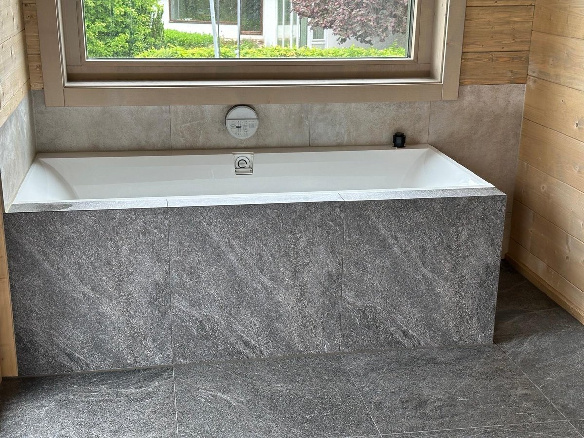 Modern bathroom with white rectangular bathtub fronted by gray stone panels, large window above, and tile flooring